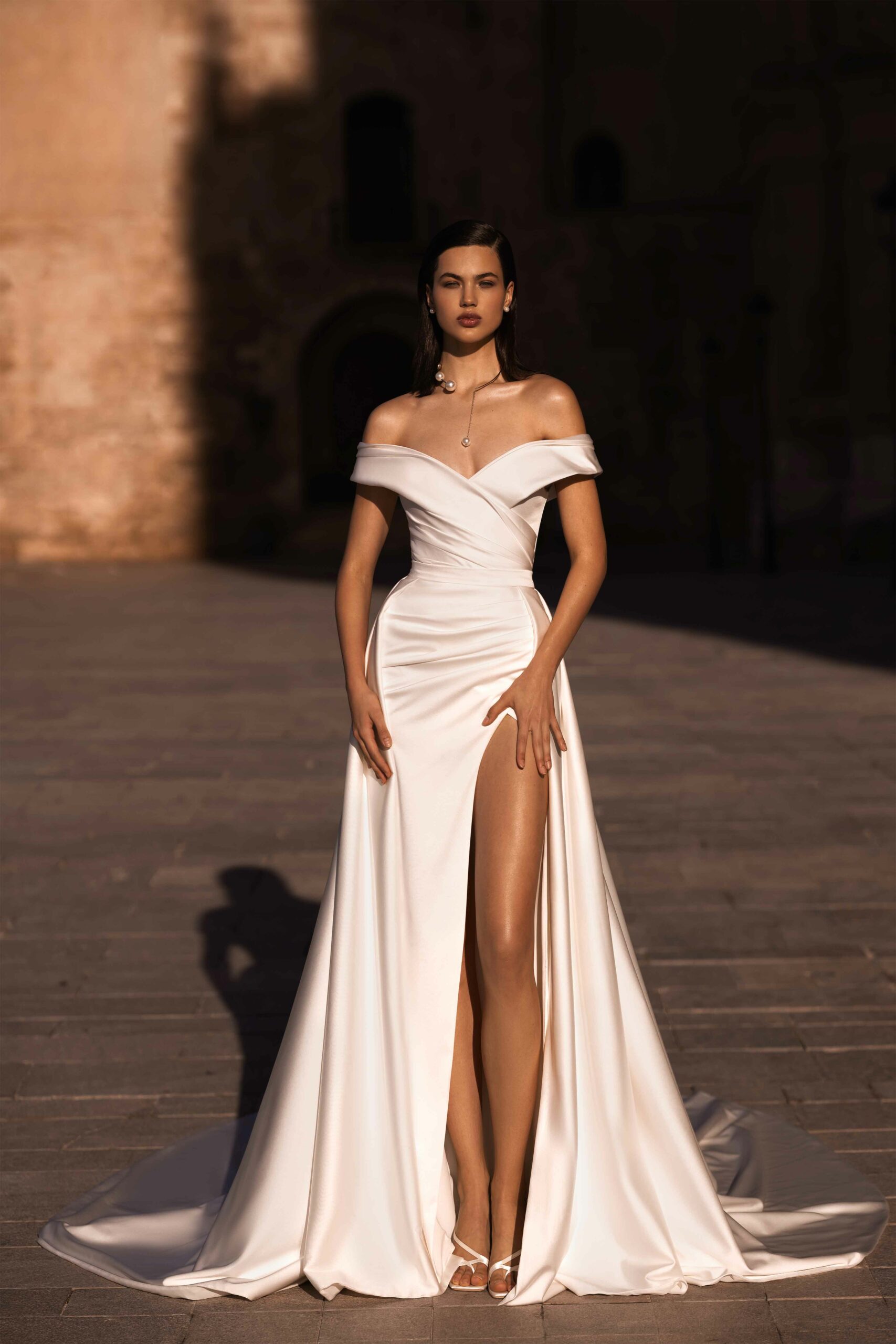 Abito da sposa Demia di Wonà con scollo off-shoulder, silhouette aderente, profondo spacco frontale e lunga gonna fluida con strascico, disponibile da Altomare Sposa a Modena.