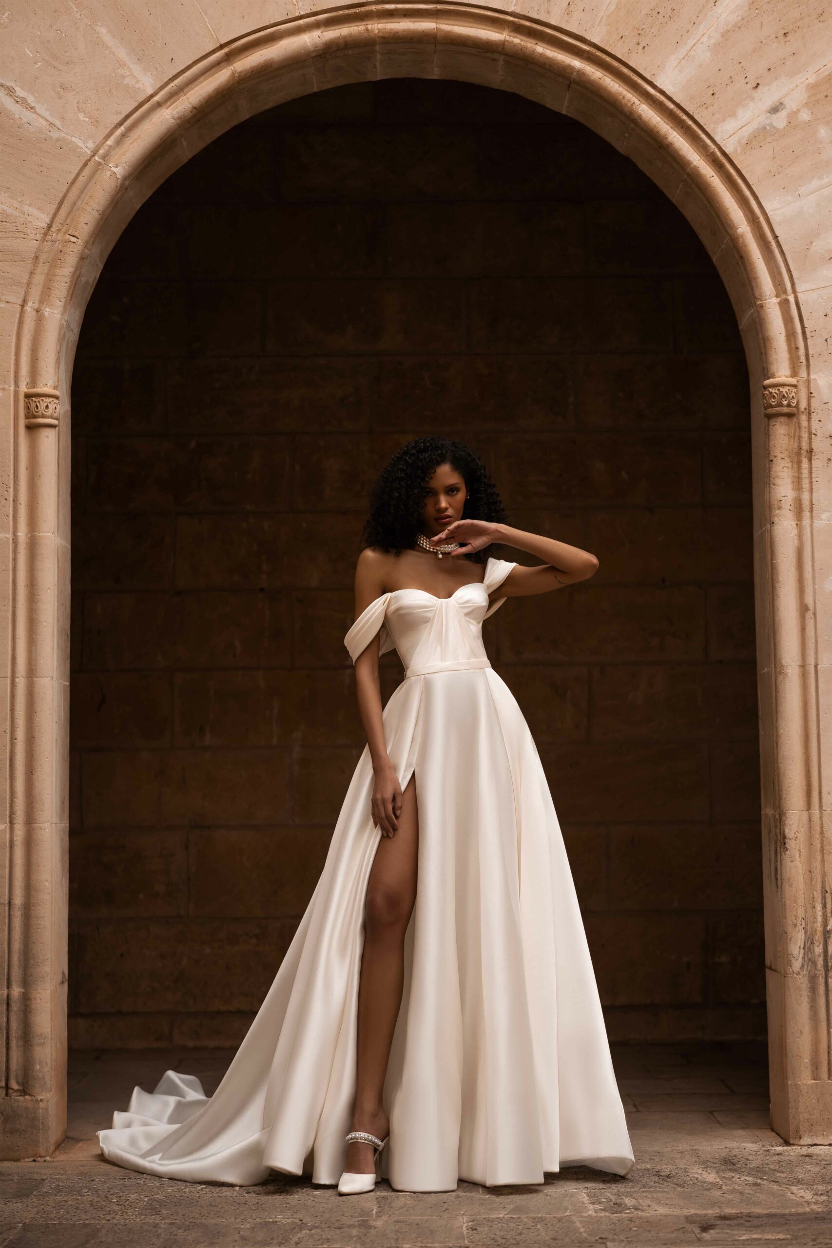 Abito da sposa Dorothy Wonà in mikado con spalle scese, corpetto drappeggiato, gonna ampia con spacco frontale e lunga coda, disponibile presso Altomare Sposa a Modena.