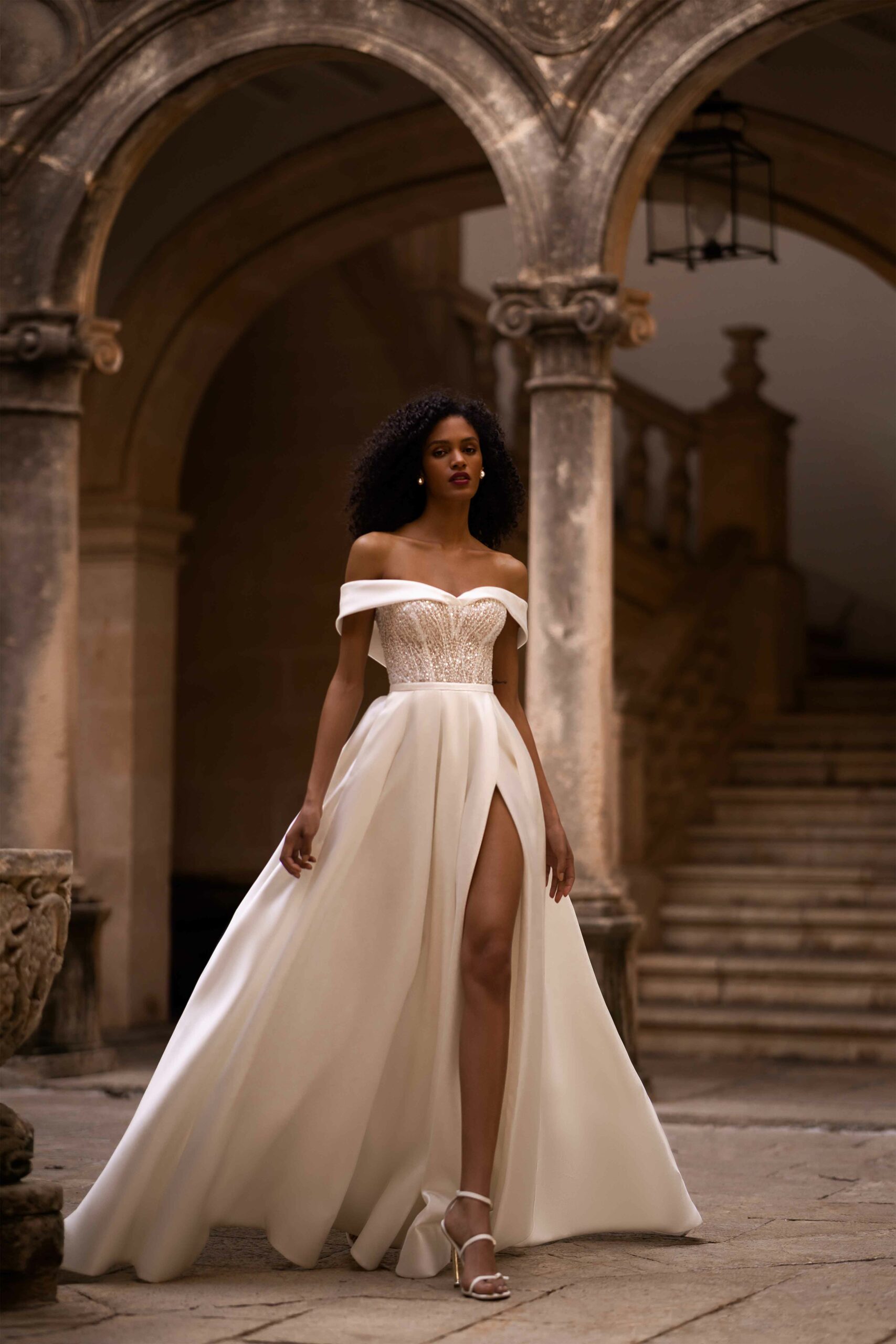 Abito da sposa Lotus di Wonà con scollo off shoulder, corpetto ricamato luminoso, ampia gonna satinata con spacco alto, indossato in un elegante cortile storico, disponibile da Altomare Sposa a Modena.