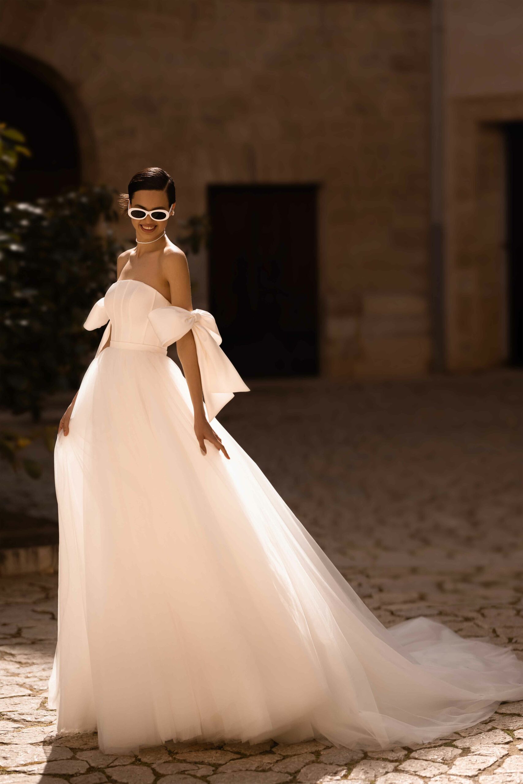 Abito da sposa Valdes di Wonà con bustier lineare, maxi fiocchi off shoulder e ampia gonna in tulle con lungo strascico, indossato in un suggestivo cortile storico, disponibile da Altomare Sposa a Modena.