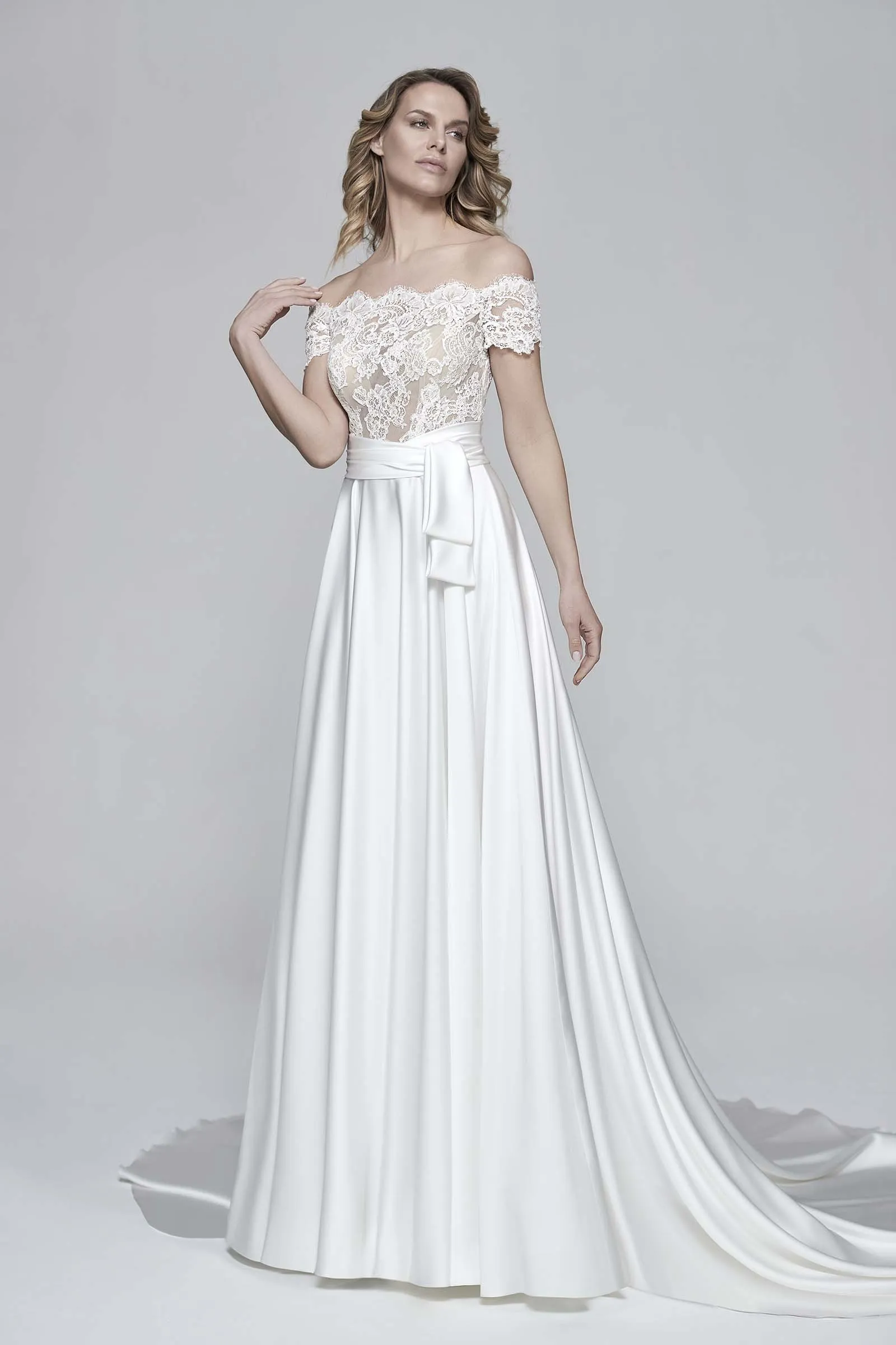 Abito da sposa Betulla di Maison Signore con scollo off shoulder in pizzo floreale, cintura in vita e gonna ampia liscia con lungo strascico, fotografato su sfondo neutro da studio.