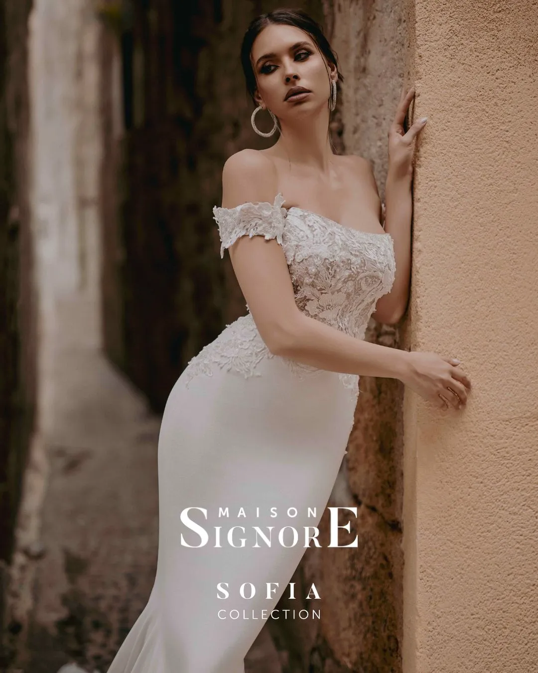 Abito da sposa Decumani di Maison Signore con linea sirena, scollo off shoulder e corpino ricamato floreale, fotografato in un suggestivo vicolo in pietra dall’atmosfera elegante e urbana.