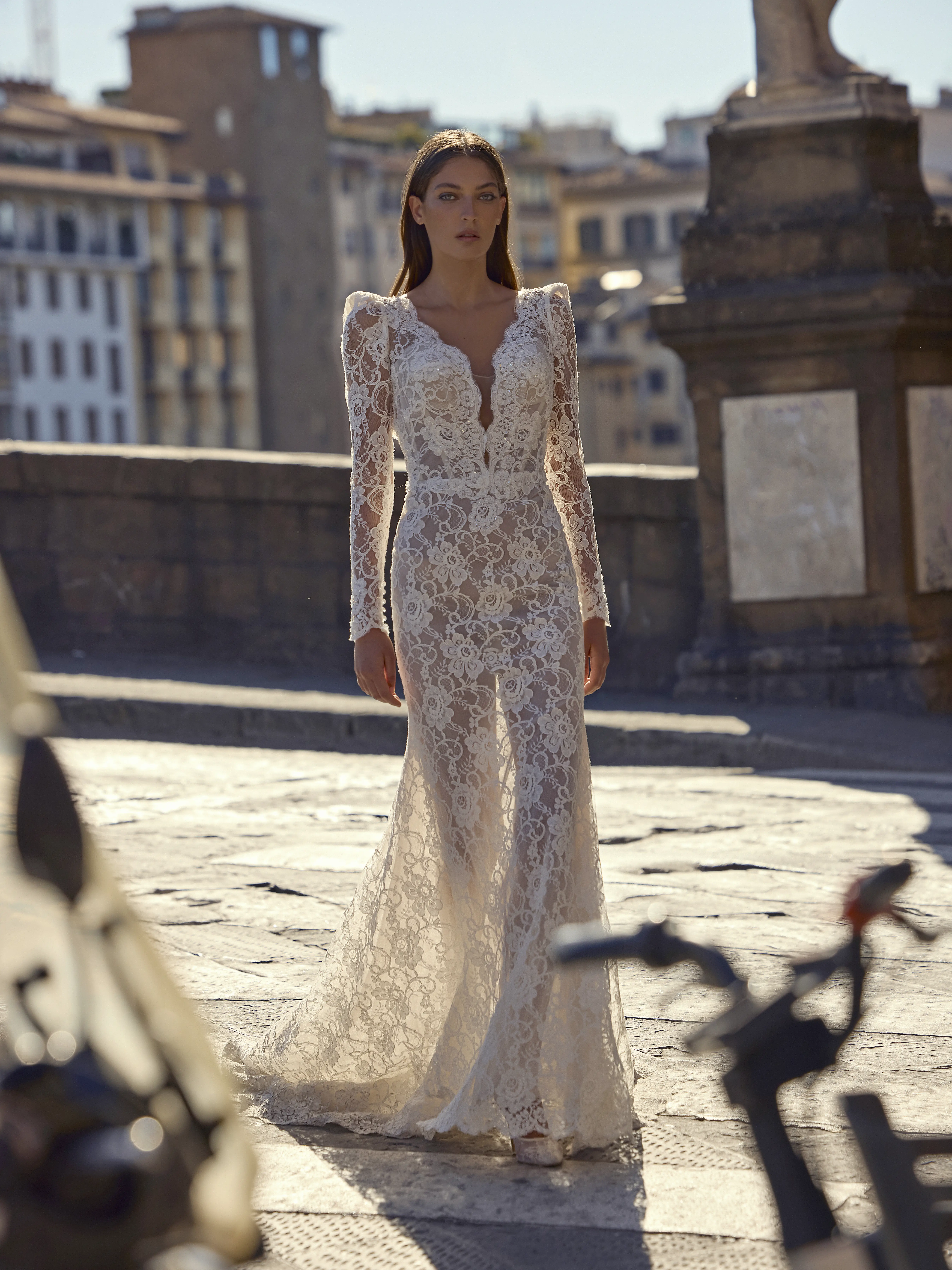 Abito da sposa sirena in pizzo con maniche lunghe e scollo profondo indossato su ponte in città storica.