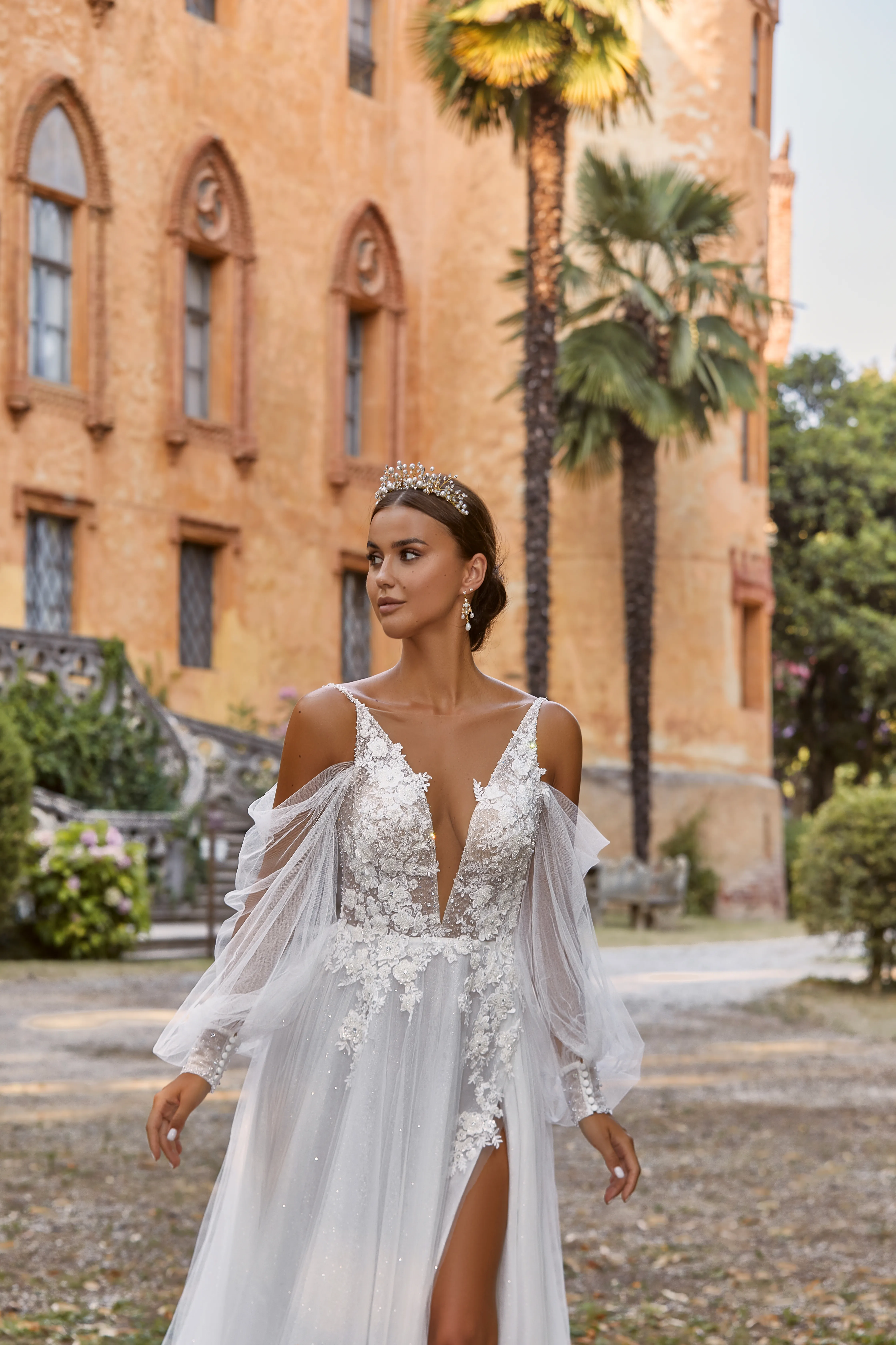 Abito da sposa Elisabetta di Maison Signore con corpetto floreale 3D, scollo a V profondo, maniche in tulle trasparente e gonna morbida con spacco, fotografato in un elegante giardino davanti a una villa storica.
