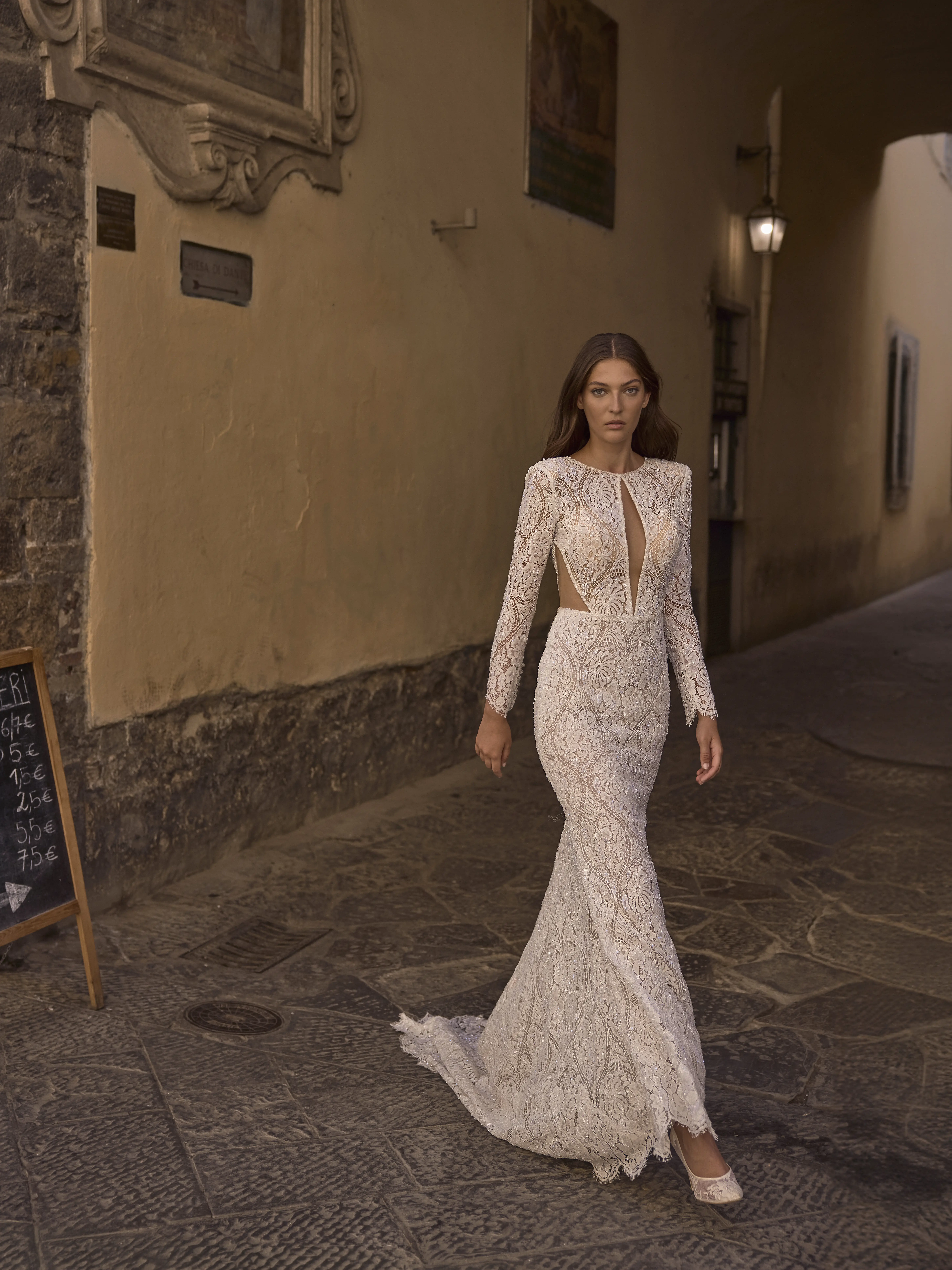 Sposa indossa abito Etherea in pizzo aderente con maniche lunghe e scollatura profonda, camminando in un vicolo elegante in stile italiano.