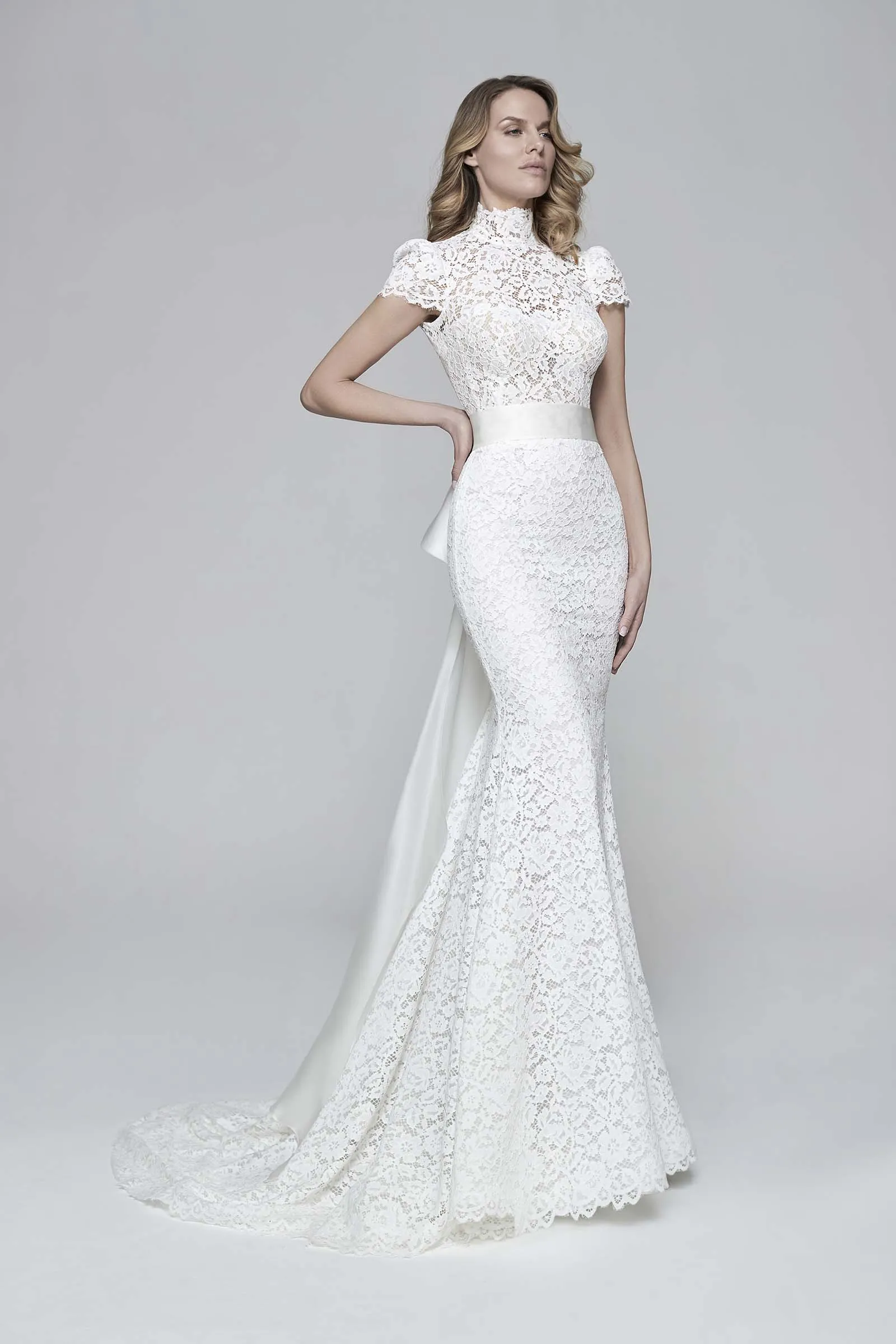 Abito da sposa Gerbera di Maison Signore con linea sirena in pizzo, collo alto, maniche corte e lungo fiocco posteriore, fotografato su sfondo neutro da studio.