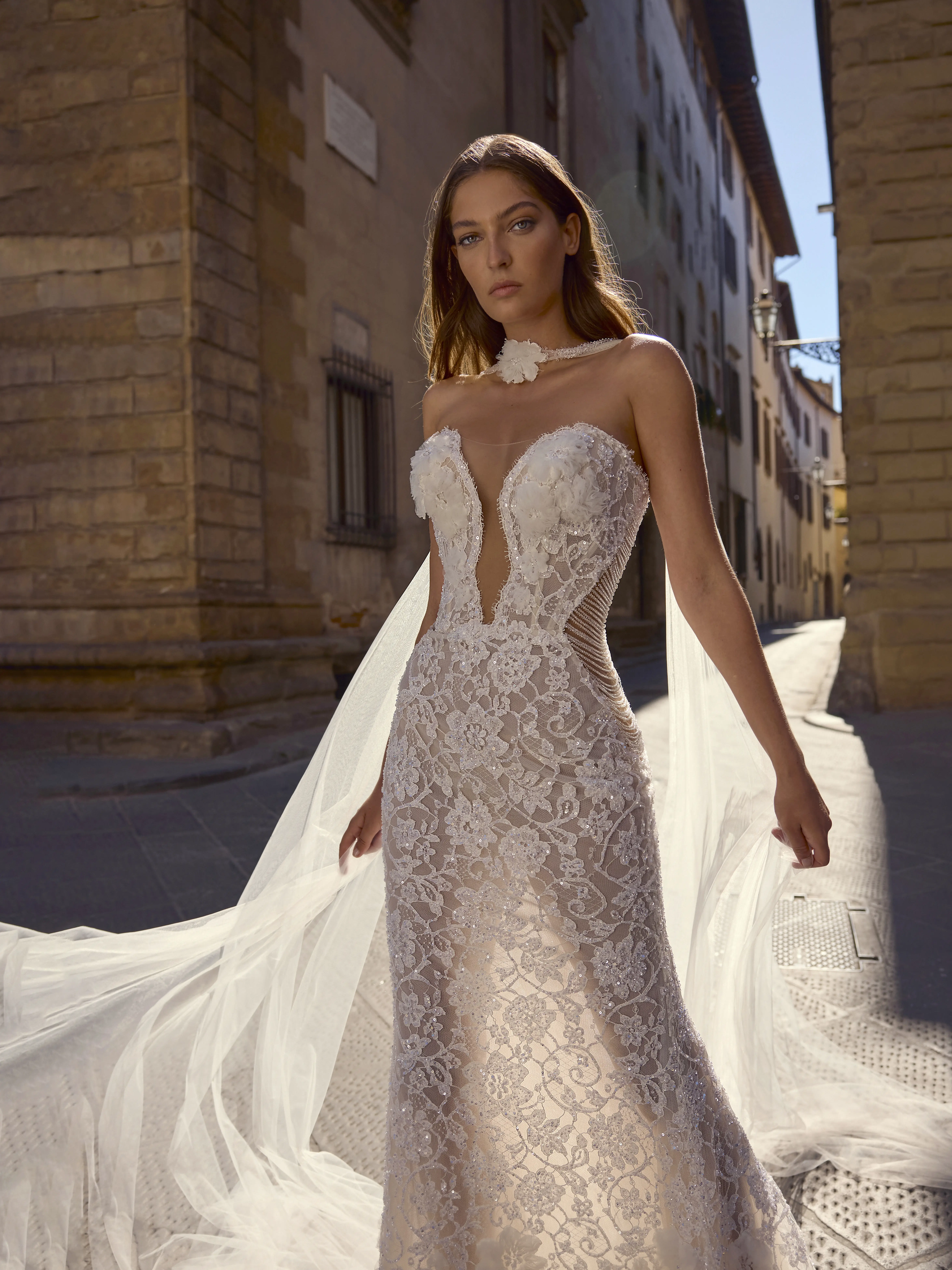 Sposa indossa abito Helios in pizzo aderente con scollatura profonda e velo fluido, camminando in una strada storica illuminata dal sole.