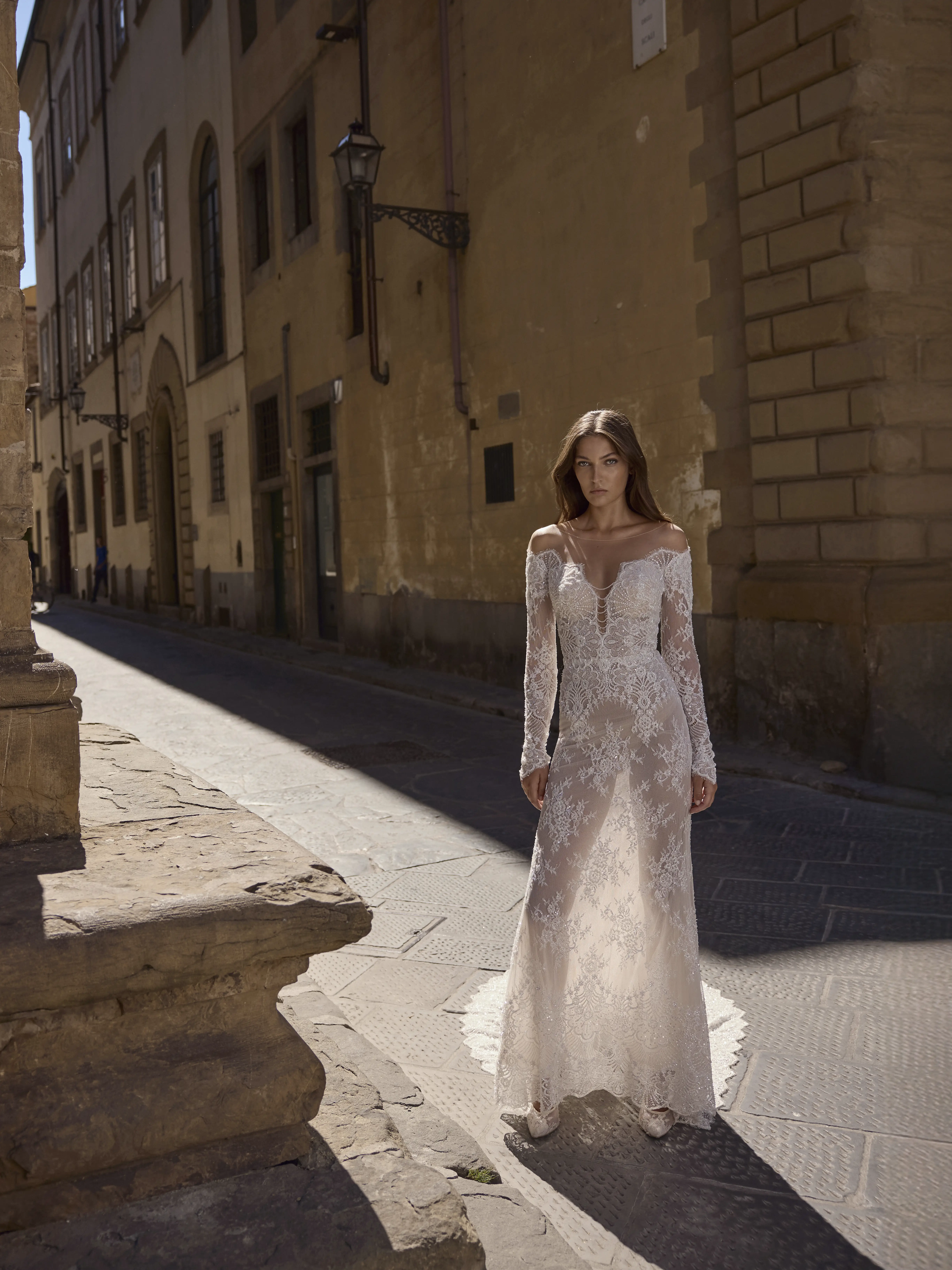 Sposa indossa abito Maia in pizzo con maniche lunghe e scollatura morbida, in piedi in una strada storica con luce naturale.