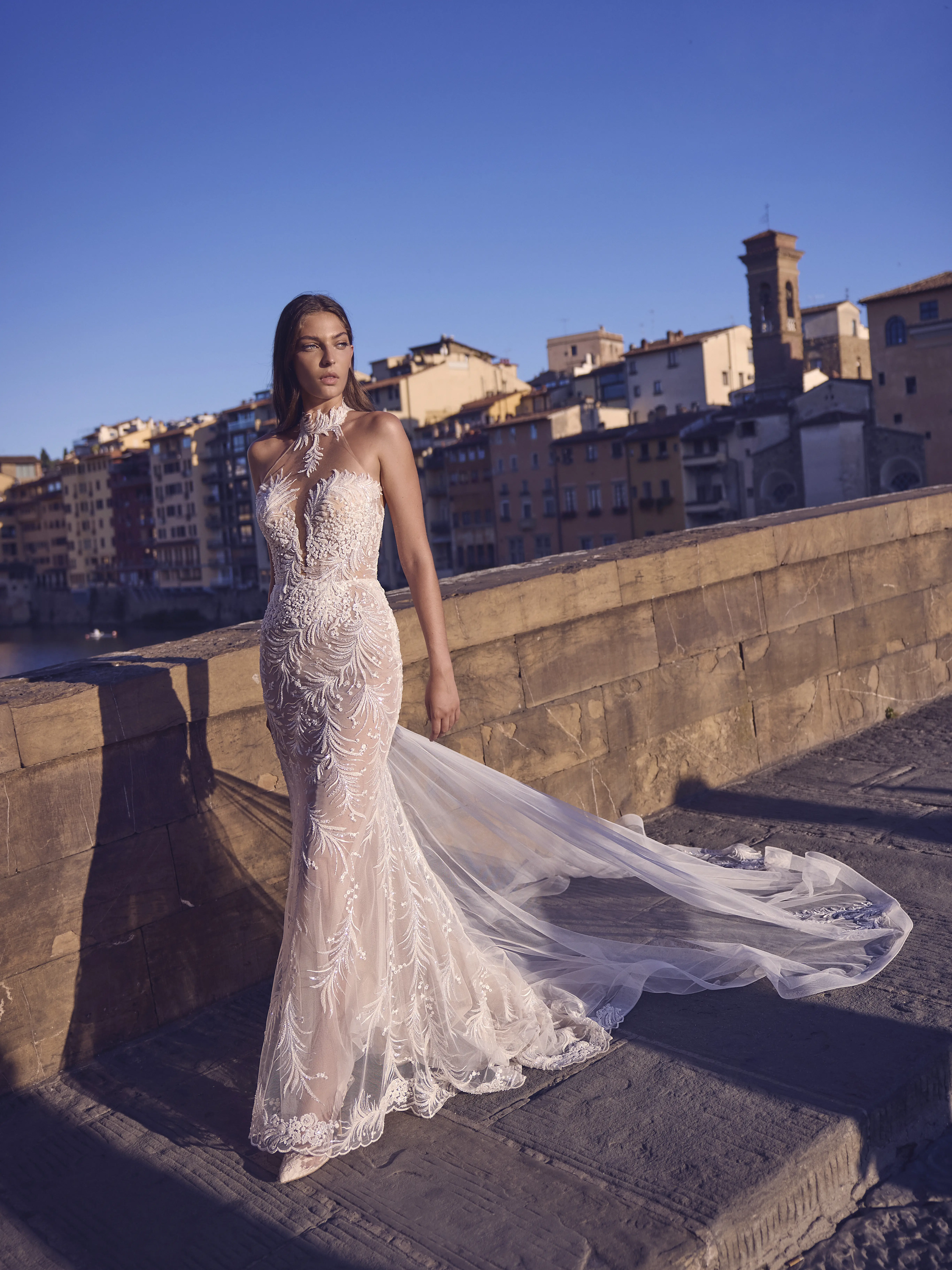 Abito da sposa sirena ricamato con collo alto e lungo strascico in tulle indossato su ponte con città storica sullo sfondo.