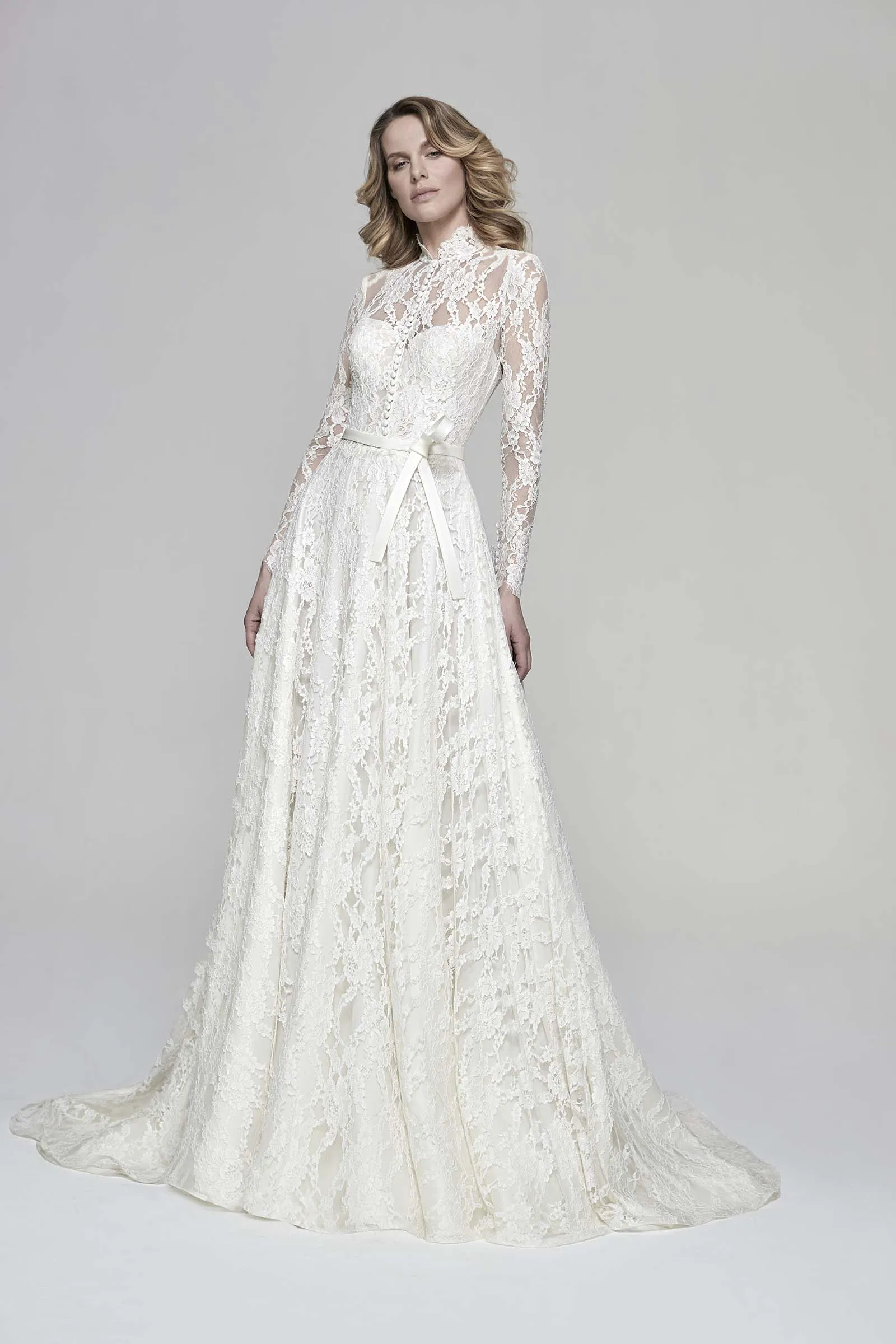 Abito da sposa Miltonia di Maison Signore in pizzo totale con collo alto, maniche lunghe trasparenti, cintura sottile in vita e gonna ampia con strascico, fotografato su sfondo neutro da studio.
