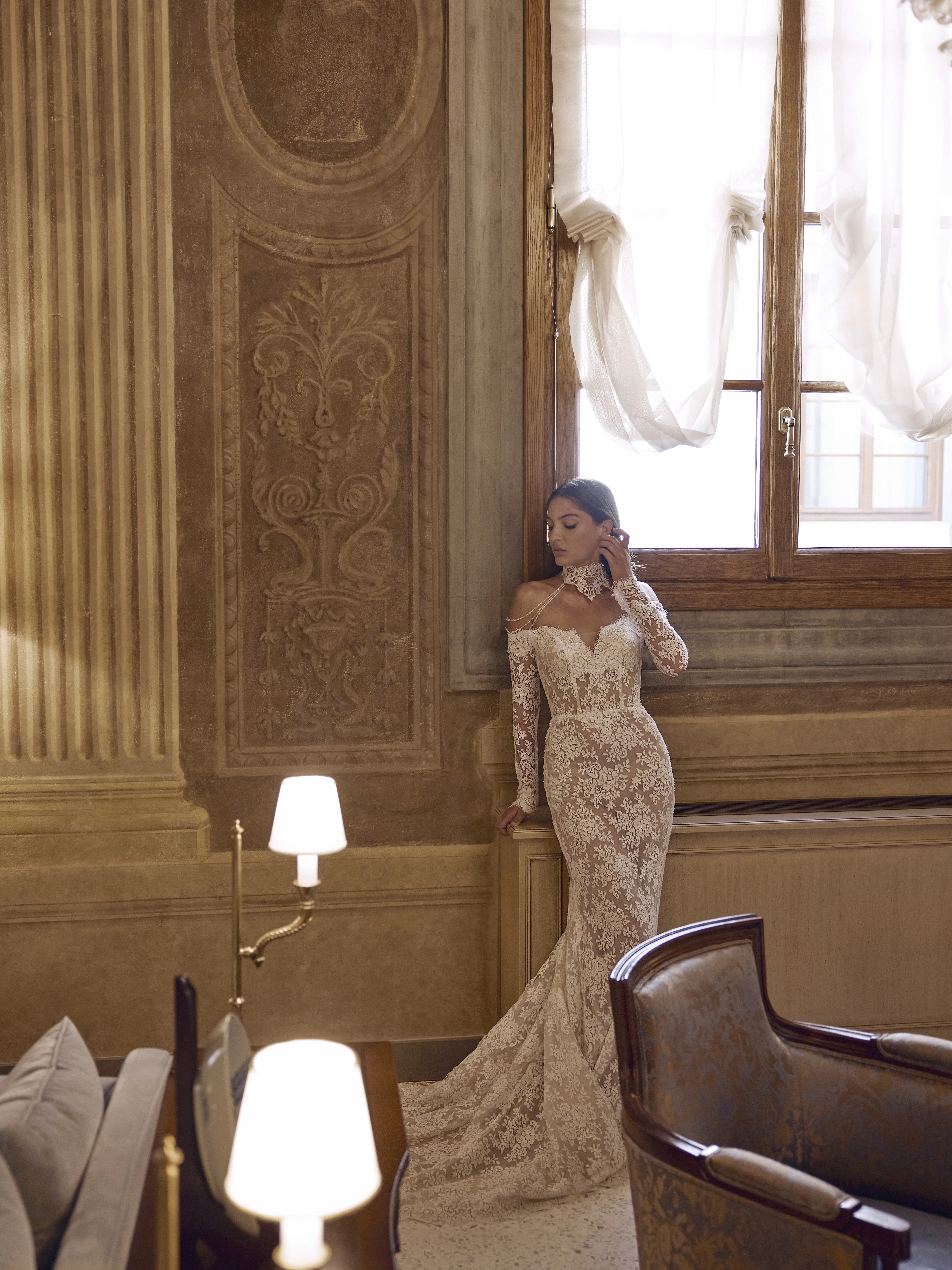 Sposa indossa abito Naiade in pizzo aderente con maniche lunghe e scollo off shoulder, in un elegante interno classico vicino a una finestra.
