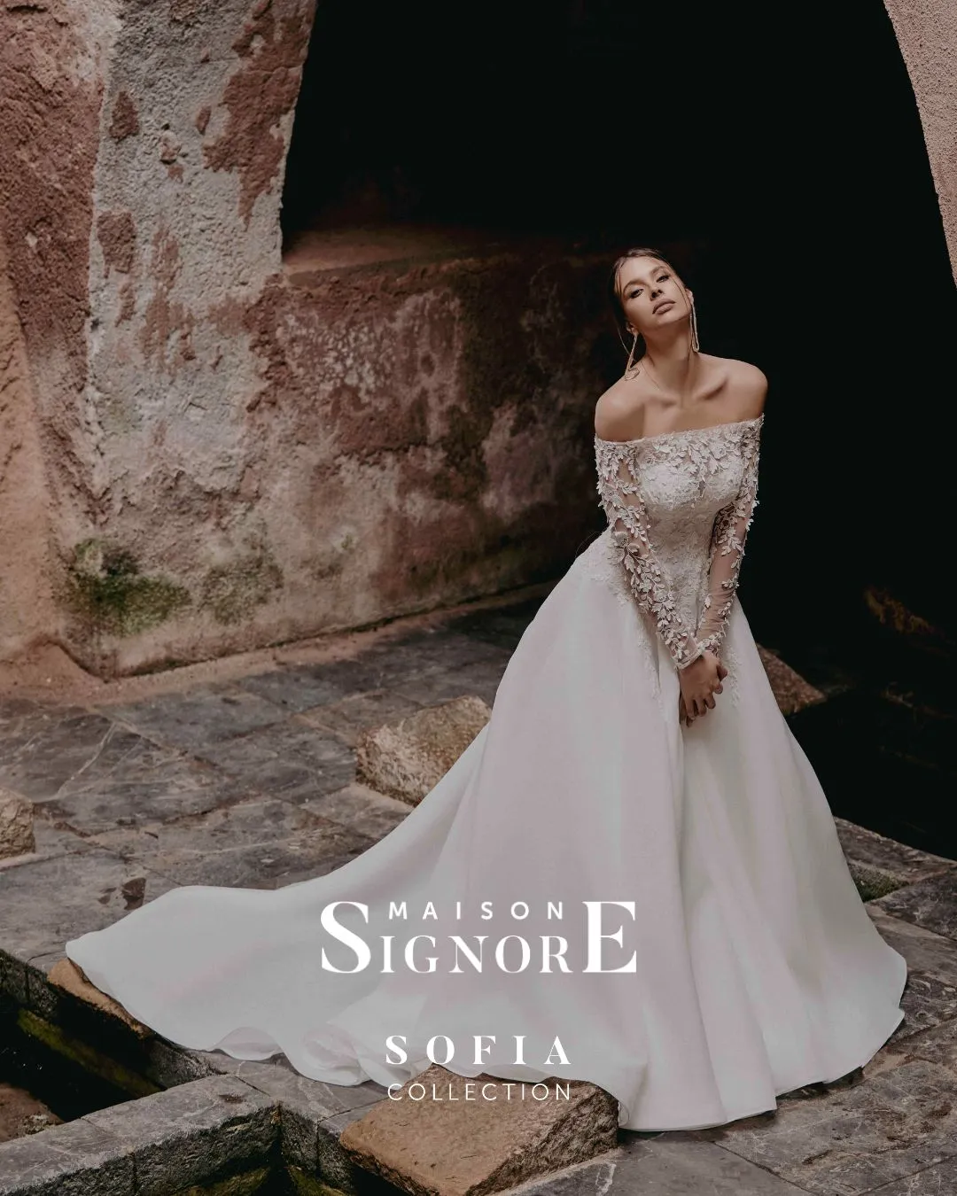 Abito da sposa Portalba di Maison Signore con scollo off shoulder, maniche lunghe in pizzo floreale tridimensionale e ampia gonna principesca, fotografato in un suggestivo contesto in pietra dal fascino storico.
