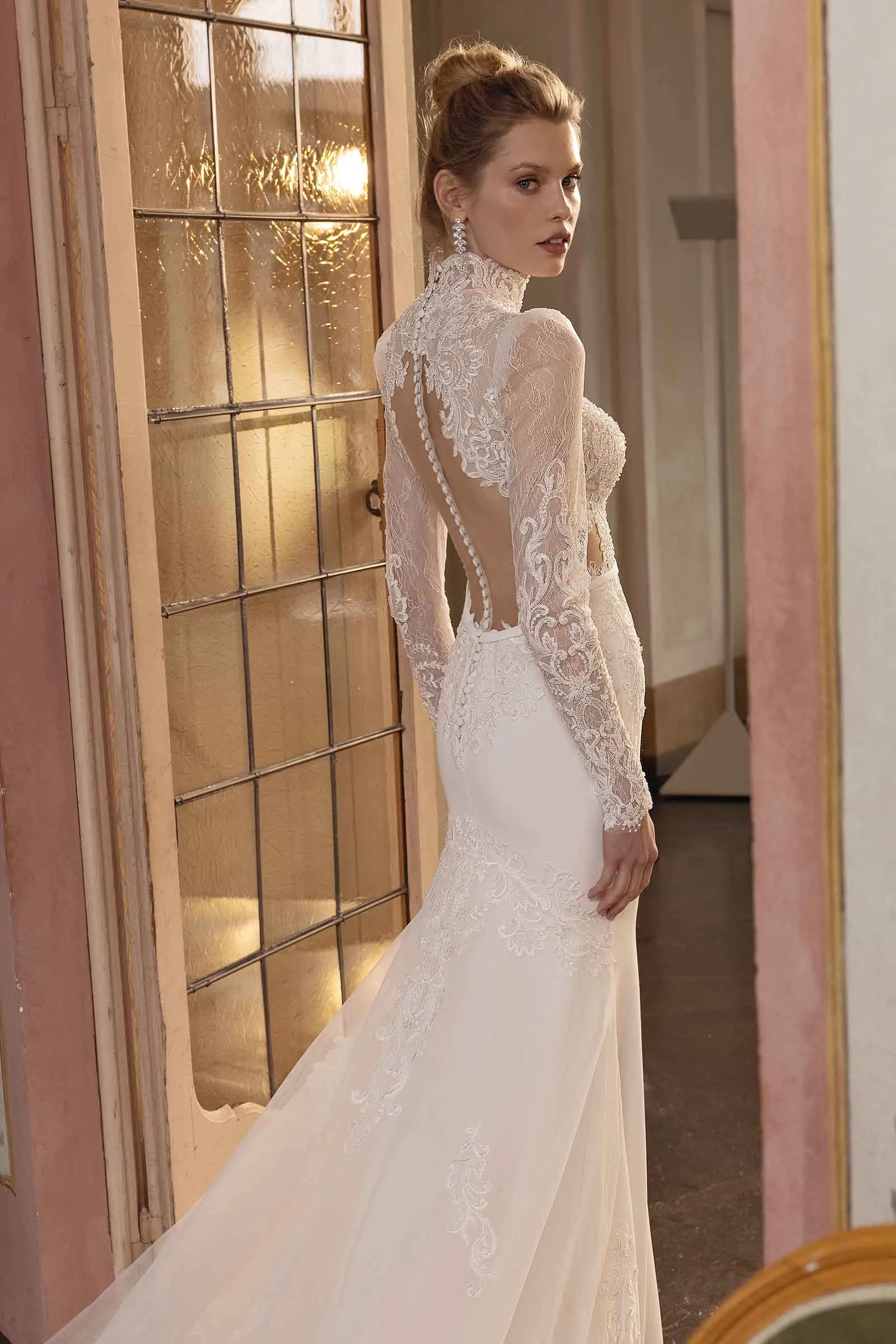 Abito da sposa Shanghai di Maison Signore con linea sirena, maniche lunghe in pizzo, collo alto e schiena scoperta effetto tattoo, fotografato in un elegante interno con porta vetrata illuminata.