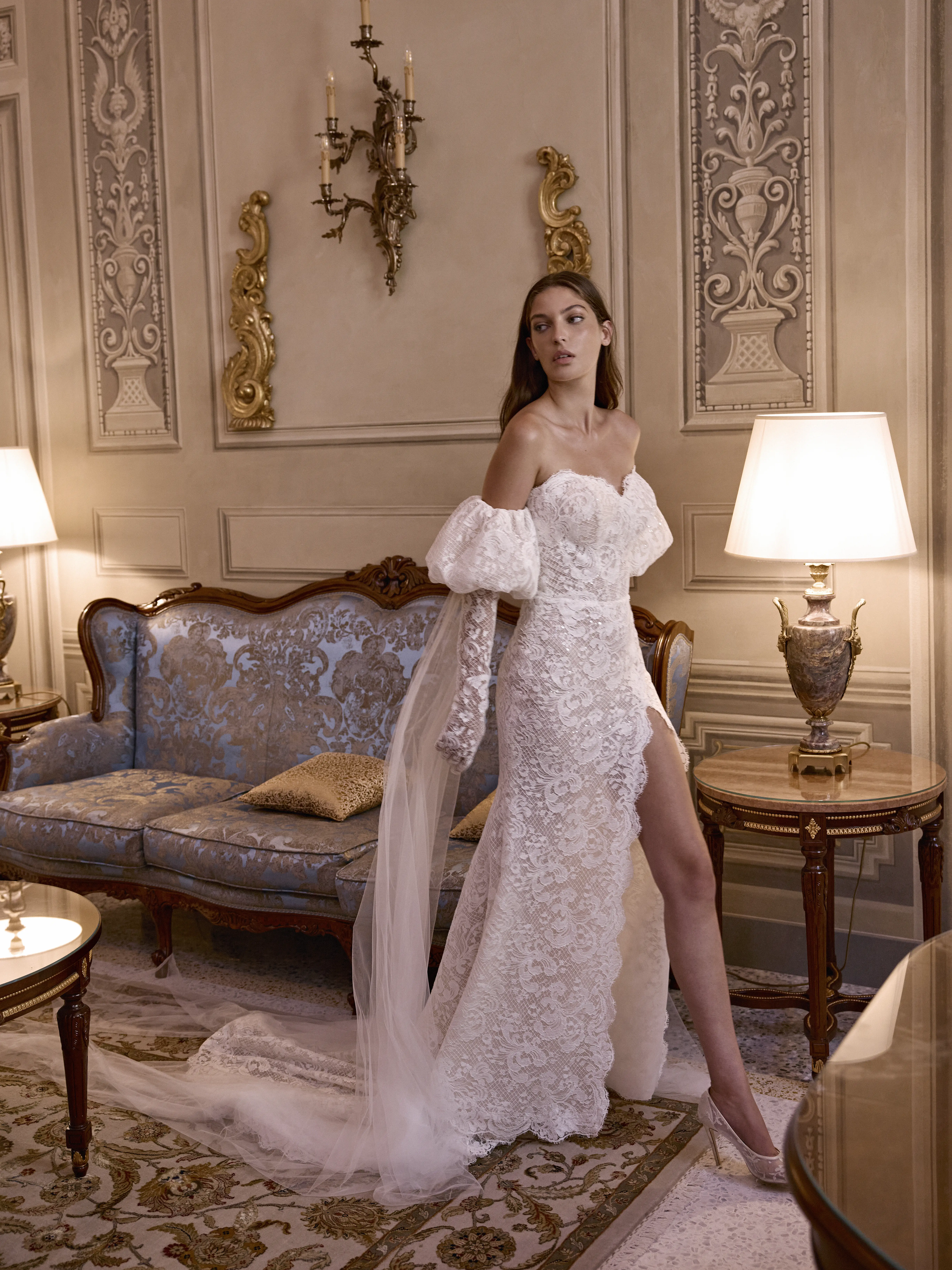 Abito da sposa sirena in pizzo con maniche off-shoulder e spacco laterale indossato in elegante salotto classico.