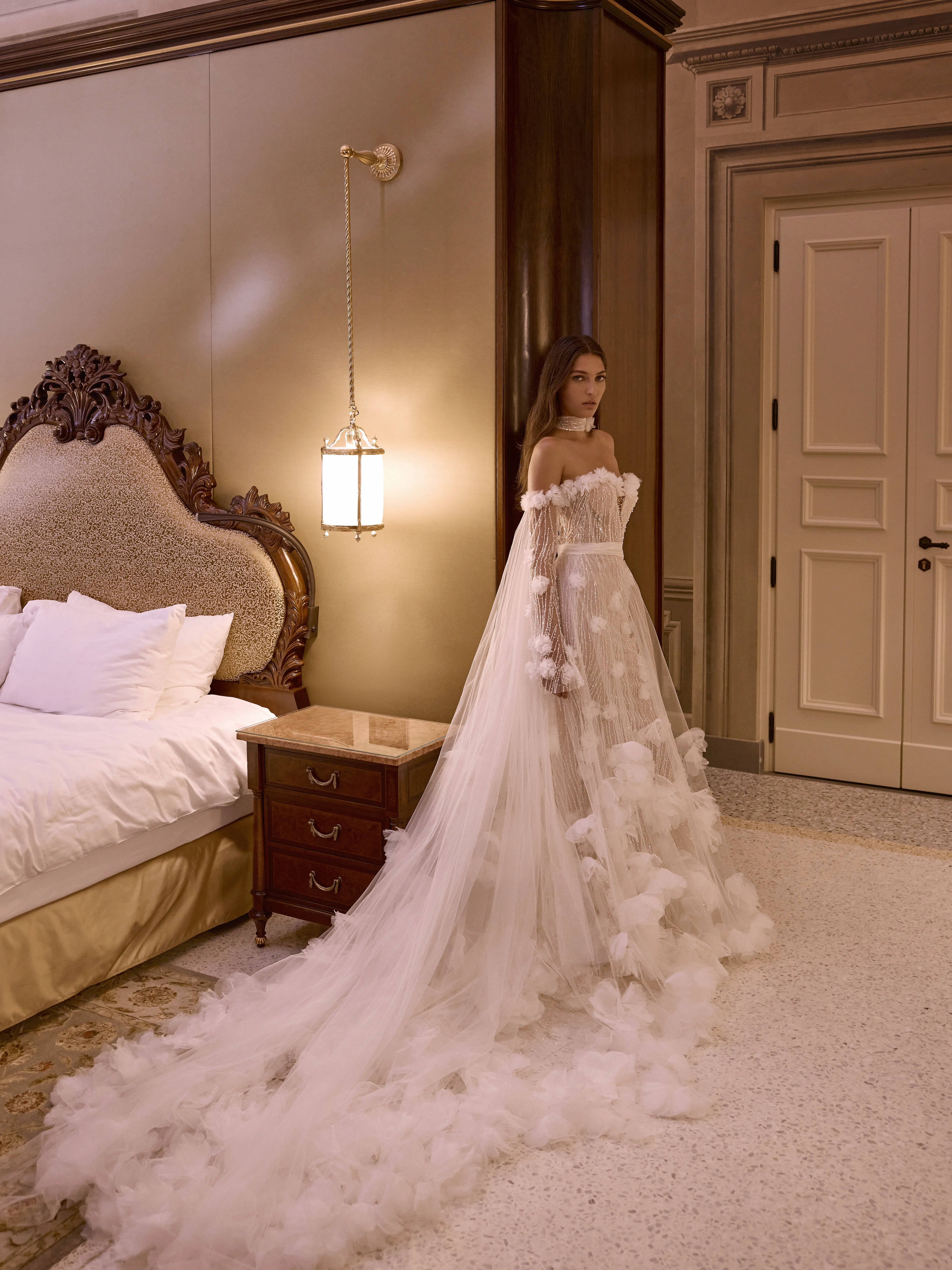 Abito da sposa con mantello in tulle e applicazioni floreali tridimensionali indossato in elegante camera con arredi classici.