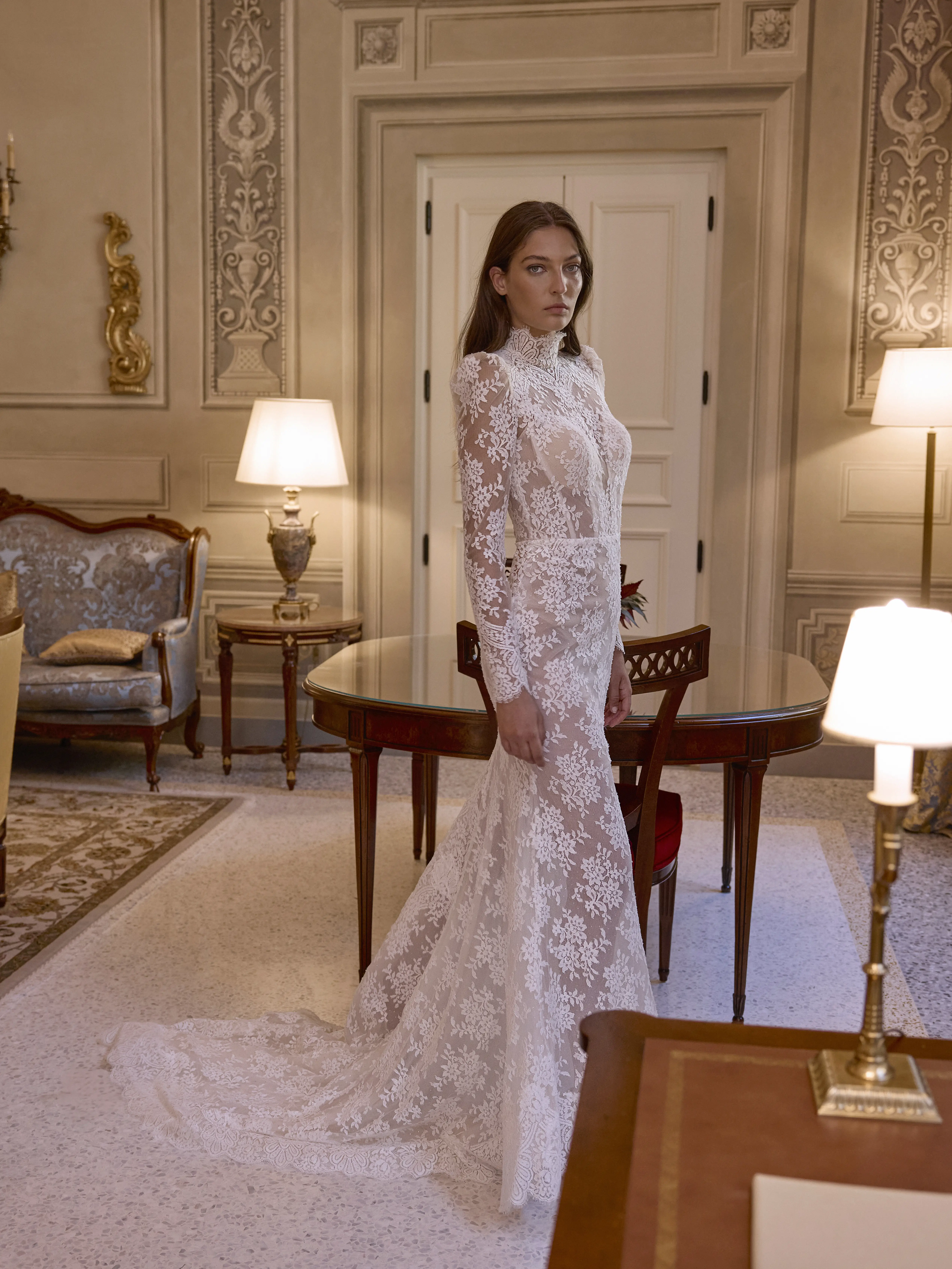 Sposa indossa abito Tritone in pizzo aderente con maniche lunghe e collo alto, in un elegante interno classico con arredi raffinati.