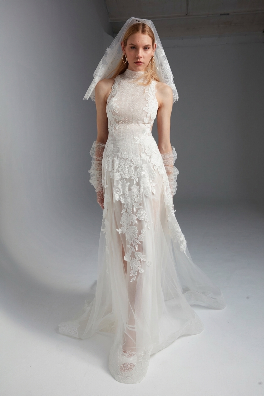 In un paesaggio costiero roccioso all'alba/tramonto, una donna indossa l'abito da sposa Eligia di Yolan cris, un abito lungo in pizzo e tulle con scollo all'americana. Lei solleva un lembo della gonna ampia, facendola fluttuare al vento e catturando la luce dorata. Il mare e le barche a vela sono visibili sullo sfondo.