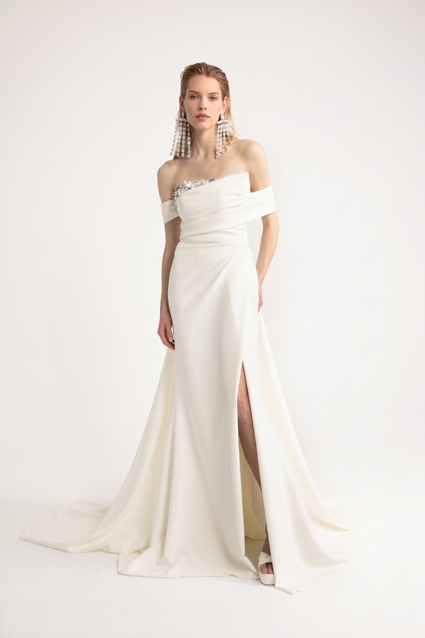 Modella indossa l'abito da sposa sofisticato Lorenza di Yolan Cris, un modello a sirena con scollo omerale, corpetto drappeggiato e spacco laterale su uno sfondo neutro.